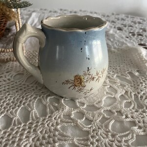 Pu&ograve; includere: Un piccolo creamer in ceramica blu e bianca con un motivo floreale e un bordo smerlato. Il creamer ha un manico ed &egrave; appoggiato su un pizzo bianco all'uncinetto.