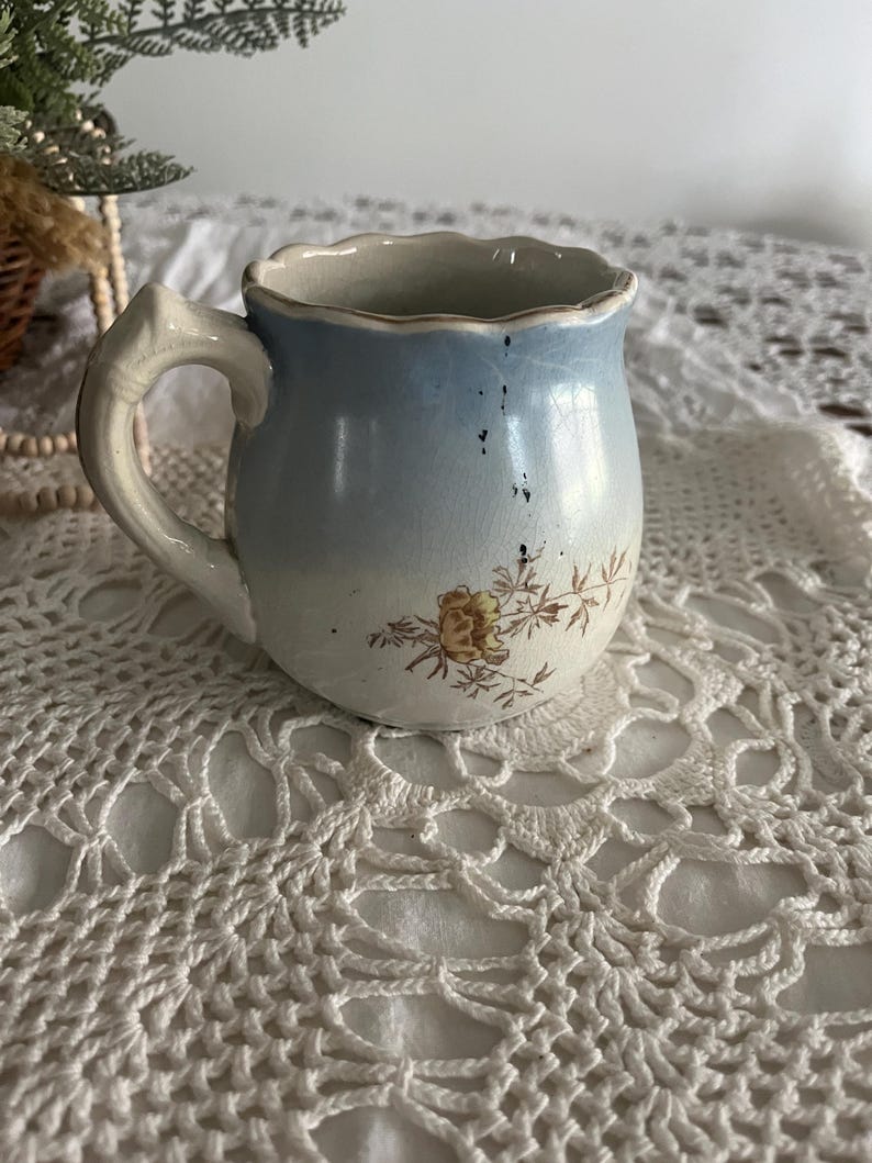 Pu&ograve; includere: Un piccolo creamer in ceramica blu e bianca con un motivo floreale e un bordo smerlato. Il creamer ha un manico ed &egrave; appoggiato su un pizzo bianco all'uncinetto.