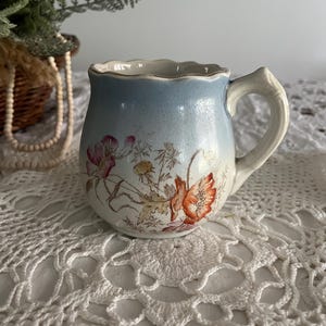 Pu&ograve; includere: Una piccola tazza in ceramica blu e bianca con un motivo floreale. La tazza ha un bordo ondulato e un manico. I fiori sono arancioni, rosa e gialli.
