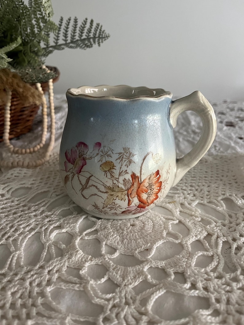 Pu&ograve; includere: Una piccola tazza in ceramica blu e bianca con un motivo floreale. La tazza ha un bordo ondulato e un manico. I fiori sono arancioni, rosa e gialli.