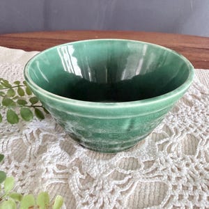 May include: A round ceramic bowl with a glossy, deep green glaze and a subtle crackle finish. The bowl features a wide, open top and a smooth, curved exterior, showcasing a consistent, vibrant emerald green color throughout the entire piece.