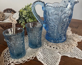 May include: A set of three blue glass pitcher and glasses with a floral pattern. The pitcher has a handle and a wide mouth. The glasses are tall and slender. The set is on a white crocheted doily on a wooden table.