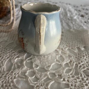 Pu&ograve; includere: Un piccolo creamer in ceramica blu e bianca con un motivo floreale e un manico dorato. Il creamer &egrave; appoggiato su un pizzo bianco all'uncinetto.