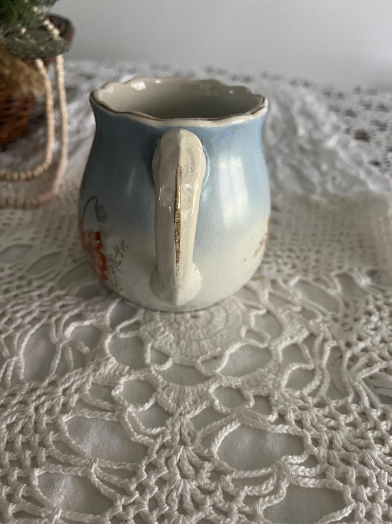 Pu&ograve; includere: Un piccolo creamer in ceramica blu e bianca con un motivo floreale e un manico dorato. Il creamer &egrave; appoggiato su un pizzo bianco all'uncinetto.