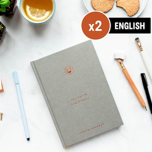 Pu&ograve; includere: Un diario grigio con il titolo "The Green Conspiracy Garden Journal" stampato in rame sulla copertina. Il diario &egrave; su una superficie bianca con una tazza di t&egrave;, una matita e una penna.