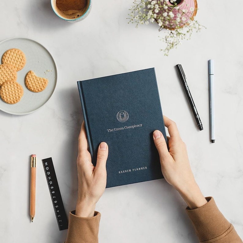 May include: A navy blue hardcover journal with the text "The Green Conspiracy" and "GARDEN PLANNER" in silver lettering. The journal is being held by a person's hands.