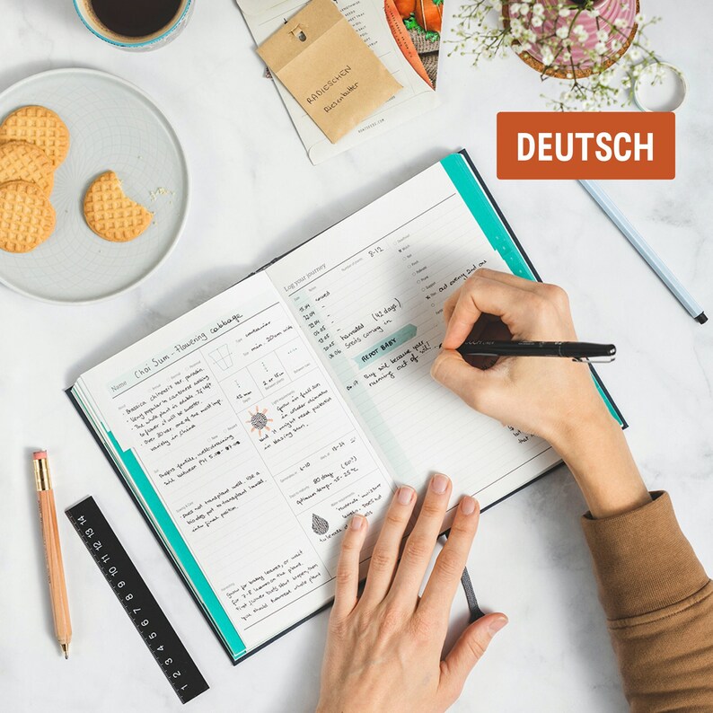 May include: A person writing in a blue and black planner with a black pen. The planner has a green tab and the words "Chai Sum Flowering cabbage" and "RADICSCHEN Riesenbuter" are visible. The planner is on a white surface with a cup of coffee, cookies, and a ruler.