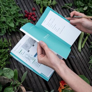 May include: A teal and white gardening journal with a monthly calendar and plant notes section. The journal is open to the September page, which lists vegetables to plant in September, including spinach, leeks, and tomatoes.