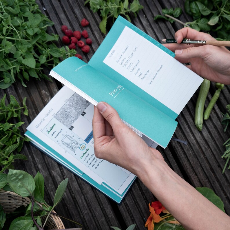 May include: A teal and white gardening journal with a monthly calendar and plant notes section. The journal is open to the September page, which lists vegetables to plant in September, including spinach, leeks, and tomatoes.