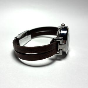 以下が含まれることがあります： 茶色の革バンドと銀色の文字盤が付いた腕時計。バンドは手首に二重に巻き付けられています。時計は白い背景に配置されています。