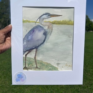 May include: A watercolor painting of a blue heron standing on a rock by the water. The heron is facing right and has its wings folded. The painting is signed "Art Gini" and dated "2023".