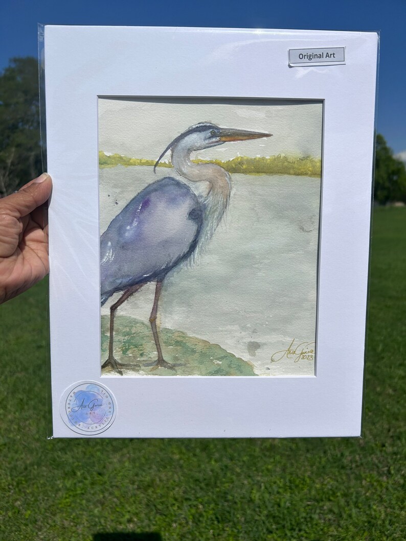 May include: A watercolor painting of a blue heron standing on a rock by the water. The heron is facing right and has its wings folded. The painting is signed "Art Gini" and dated "2023".