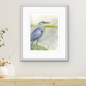 May include: A watercolor painting of a blue heron standing on a rock by a body of water. The heron is facing to the right and has its head turned to the left. The background is a light blue and green.