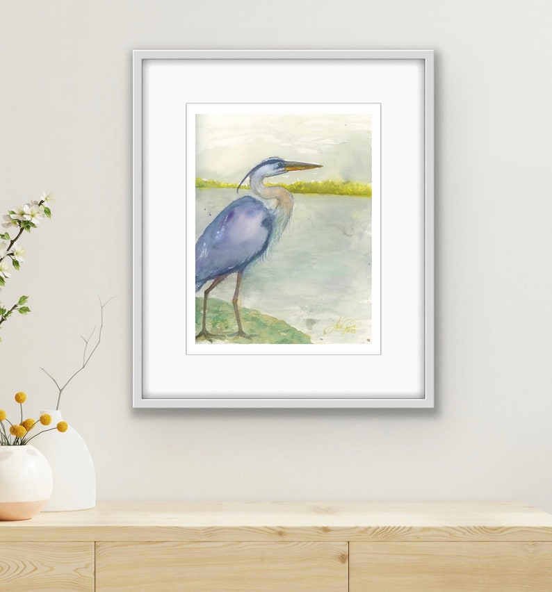 May include: A watercolor painting of a blue heron standing on a rock by a body of water. The heron is facing to the right and has its head turned to the left. The background is a light blue and green.