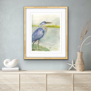 May include: A watercolor painting of a blue heron standing on a green grassy bank by a body of water. The heron is facing to the right and has its head turned to the left. The background is a light blue and green watercolor wash.