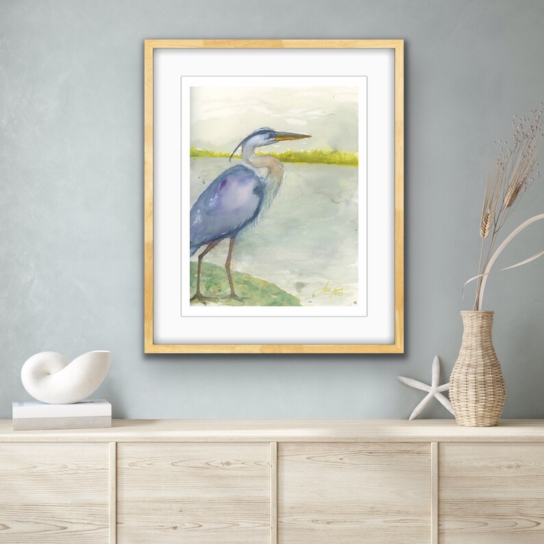 May include: A watercolor painting of a blue heron standing on a green grassy bank by a body of water. The heron is facing to the right and has its head turned to the left. The background is a light blue and green watercolor wash.
