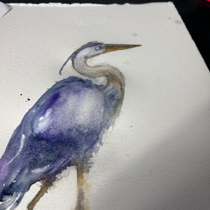 May include: Watercolor painting of a blue heron standing on one leg with its head turned to the side. The bird is painted in shades of blue and purple, with a hint of brown on its legs.