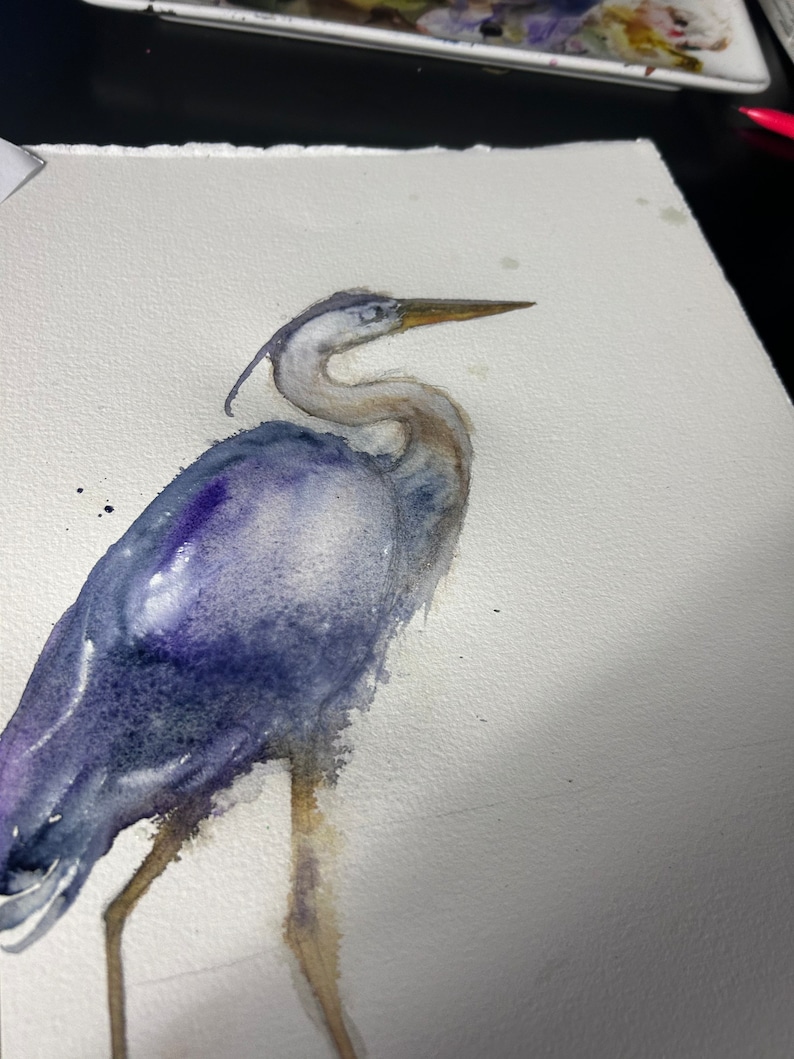 May include: Watercolor painting of a blue heron standing on one leg with its head turned to the side. The bird is painted in shades of blue and purple, with a hint of brown on its legs.