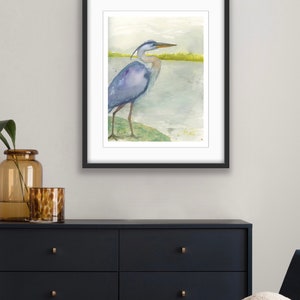 May include: Watercolor painting of a blue heron standing on a green bank by a body of water. The heron is facing to the right and has its head turned to the left. The background is a light blue and green watercolor wash.