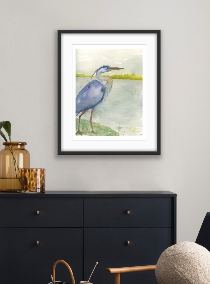 May include: Watercolor painting of a blue heron standing on a green bank by a body of water. The heron is facing to the right and has its head turned to the left. The background is a light blue and green watercolor wash.