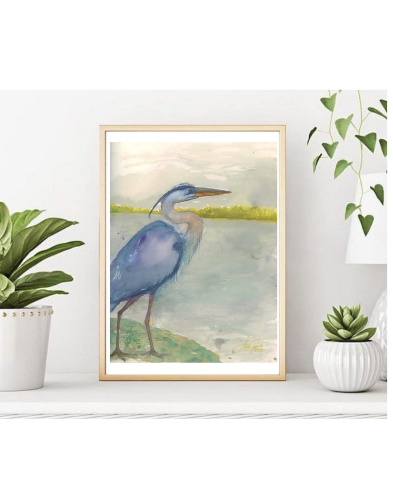 May include: A watercolor painting of a blue heron standing on a green rock by a body of water. The heron has a long neck and a long beak. The background is a soft blue and green.