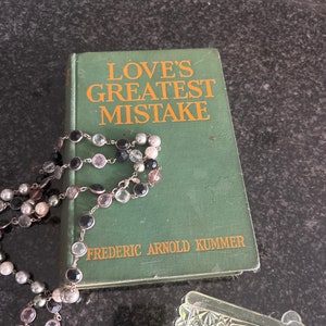 May include: A green hardback book titled "Love's Greatest Mistake" by Frederic Arnold Kummer. A beaded necklace with black, white and pink beads is draped over the book.