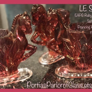 May include: Set of three ruby flashed glass vases in the shape of prancing horses. The vases are clear glass with a ruby red overlay. The vases are marked "LE Smith EAPG Ruby Flashed Glass Set of 3 Prancing Horse Vases 1910".