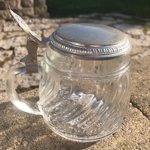 Könnte beinhalten: Ein klares Glasbierkrug mit einem silbernen Deckel und einem Scharniergriff. Der Krug hat ein strukturiertes, wellenförmiges Design.