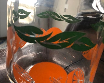 May include: A clear glass pitcher with orange and green fruit designs. The pitcher features orange citrus fruits and green leaves around the base. The pitcher has a wide base and a handle.