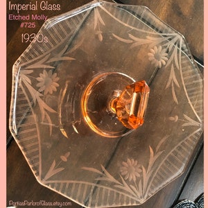 Op de afbeelding: Een glazen taartplateau met een gegraveerd bloemenpatroon en een roze glazen handvat. Het plateau is achthoekig en heeft een verhoogde rand. De tekst "Imperial Glass Etched Molly #725 1930s" is zichtbaar in de afbeelding.