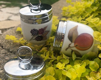 May include: Two silver egg cups with lids, one with a blackberries and one with a red fruit design. The egg cups are on a green leafy surface.