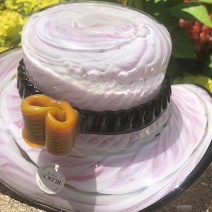 Pode incluir: Um chapéu de vidro branco e roxo com uma faixa preta e um laço amarelo. O chapéu é soprado na boca e polido. O rótulo diz "Block Crystal, Mouth Blown, Polished".