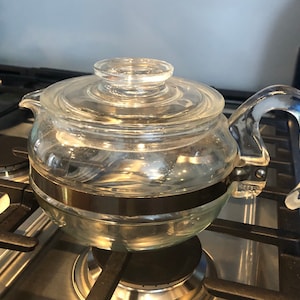 Puede incluir: Una cafetera de vidrio transparente con una banda de metal alrededor del medio. La cafetera está sobre una estufa negra.