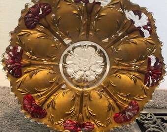 以下が含まれることがあります： 金色の縁取りと赤と白のカーネーションの花がついた透明なガラス製のサービングディッシュ。ディッシュは金色の金属製のスタンドの上に置かれています。