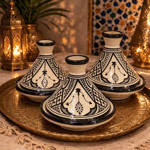 Peut inclure: Trois tajines en céramique avec des motifs géométriques noirs et blancs sont présentés sur un plateau doré. Les tajines ont des couvercles coniques et des bases circulaires. L'arrière-plan comprend des lanternes décoratives, créant une ambiance chaleureuse.