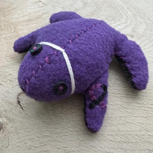 May include: A purple plush toy shaped like a sea creature with black button eyes and white stitching. The toy has a white string tied around its body.