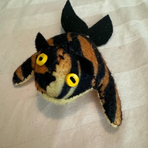 Puede incluir: Un juguete de peluche hecho a mano con forma de pez con un patrón a rayas en negro, marrón y crema. El pez tiene ojos de botón amarillos y aletas negras. El juguete está hecho de tela suave y está diseñado para ser tierno.