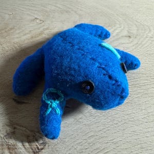 以下が含まれることがあります： 手作りの青いフェルトの生き物で、ボタンの目とステッチのディテールが施されています。 ぬいぐるみは背中に水色のアクセントがあり、手足の1つに小さなリボンが付いています。 おもちゃは木製の表面にあります。