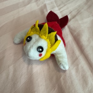 Puede incluir: Un peluche de pez koi de fieltro hecho a mano. El peluche presenta un cuerpo blanco, una espalda roja y una corona amarilla. Tiene ojos de botón negros y una mejilla roja. El pez está sobre una superficie rosa.