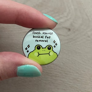 May include: A round button featuring a green frog cartoon and the text "Frogs against buccal fat removal" in black.