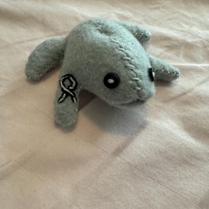 May include: A handmade gray felt creature with button eyes and stitched details. The plush toy has four appendages and a black and white ribbon emblem on one side. The toy is lying on a pink fabric surface.