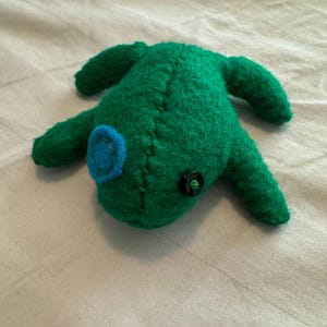 May include: A handmade green felt creature with four limbs and a rounded body. It has a blue circular detail on its face and a black button eye. The creature is stitched together with visible thread.