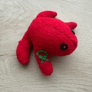 May include: A handmade red felt creature with a button eye and embroidered green detail. The plush toy has a rounded body with two small arms and a tail. The toy is made of a soft, textured material.