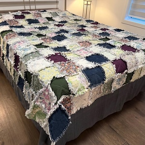May include: A patchwork quilt in shades of navy, green, purple, and floral designs covers a bed. The quilt features a fringed edge and is draped over a grey bed skirt. The bed is made up.