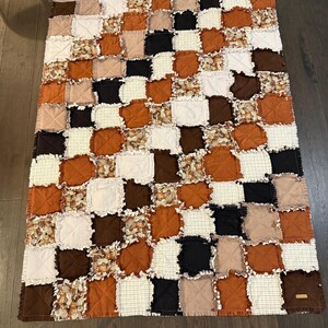 Peut inclure: Une courtepointe patchwork faite à la main avec un motif quadrillé. Elle présente des carrés de tissus blancs, noirs, bruns, beiges et oranges. Certains carrés ont un motif floral. Les bords sont effilochés.