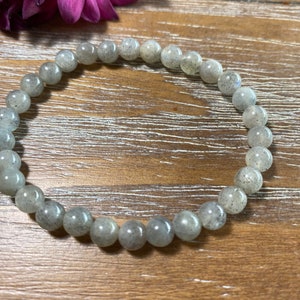 May include: A beaded bracelet made of light gray, round gemstones. The bracelet is resting on a wooden surface, with a purple flower in the background. The gemstones have a speckled appearance.
