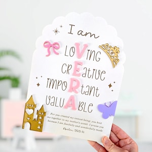 Puede incluir: Tarjeta blanca con bordes festoneados, con las palabras "I am Loving, Creative, Important, Valuable." Adornada con un lazo rosa, una corona dorada, una mariposa morada y un castillo dorado. Incluye Salmo 139:13-14.