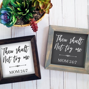 以下が含まれることがあります： 白い背景と黒い背景に「Thou shalt Not try me MOM 24:7」という文字が書かれた額縁付きの看板2つ。看板は白い木製の背景の上に置かれている。