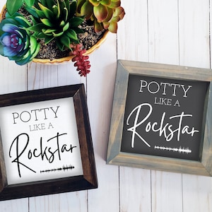 May include: Two wooden framed signs with the text "Potty Like A Rockstar" in white lettering on a black background. The sign on the left has a white sound wave graphic below the text. The sign on the right has a gray sound wave graphic below the text.