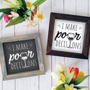 May include: Two framed signs with the text "I make poor decisions" and a glass of wine illustration. The signs are on a white wood background with flowers.